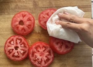 hand with a paper towel patting tomato slices dry on a cutting board.