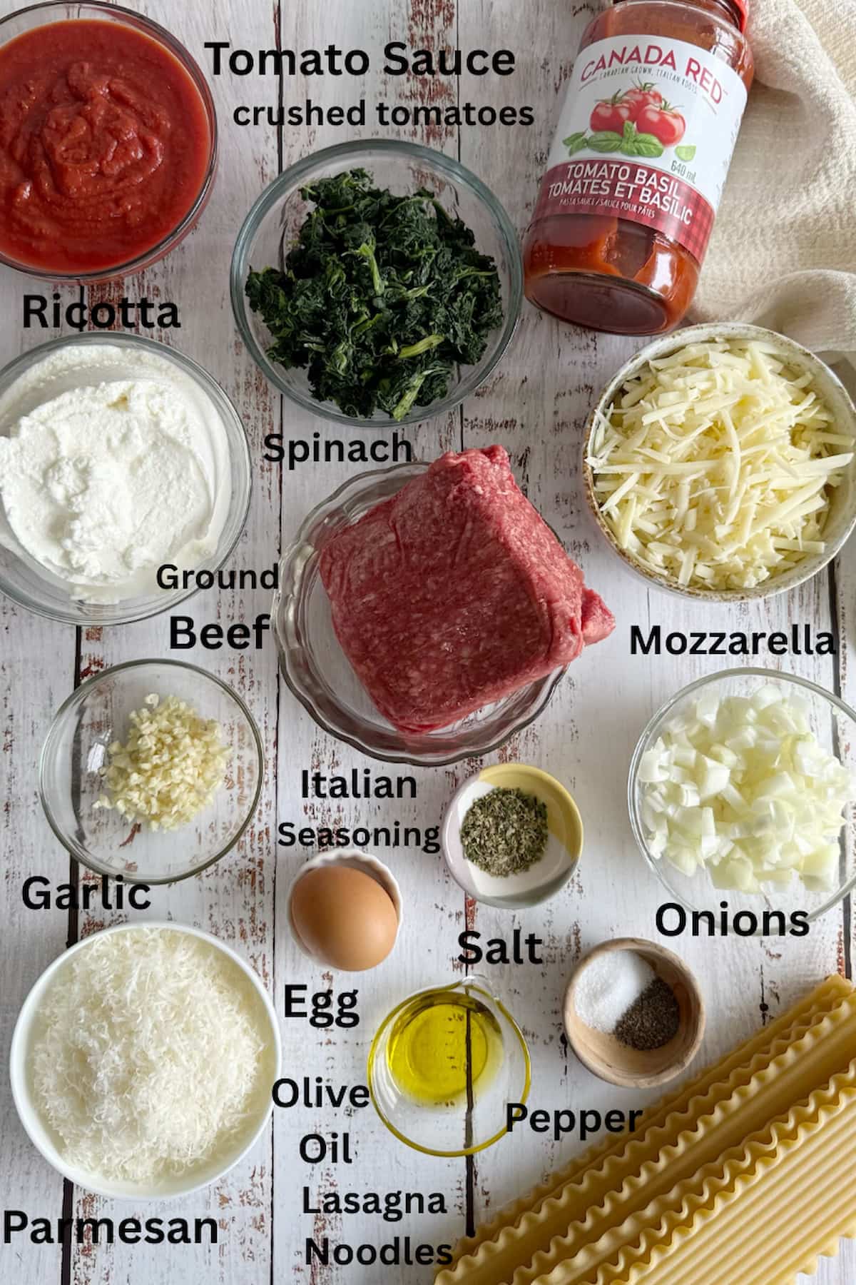 ingredients for beef and spinach lasagna laid out in separate bowls - tomato sauce, crushed tomatoes, ground beef, mozzarella, ricotta, spinach, garlic, onions, italian seasoning, egg, olive oil, lasagna noodles, salt, pepper, and parmesan cheese.