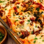 Close up of spinach and ground beef lasagna in a glass baking dish, topped with melted cheese, savoury meat sauce, and fresh parsley, with bowls of herbs and red pepper flakes nearby.