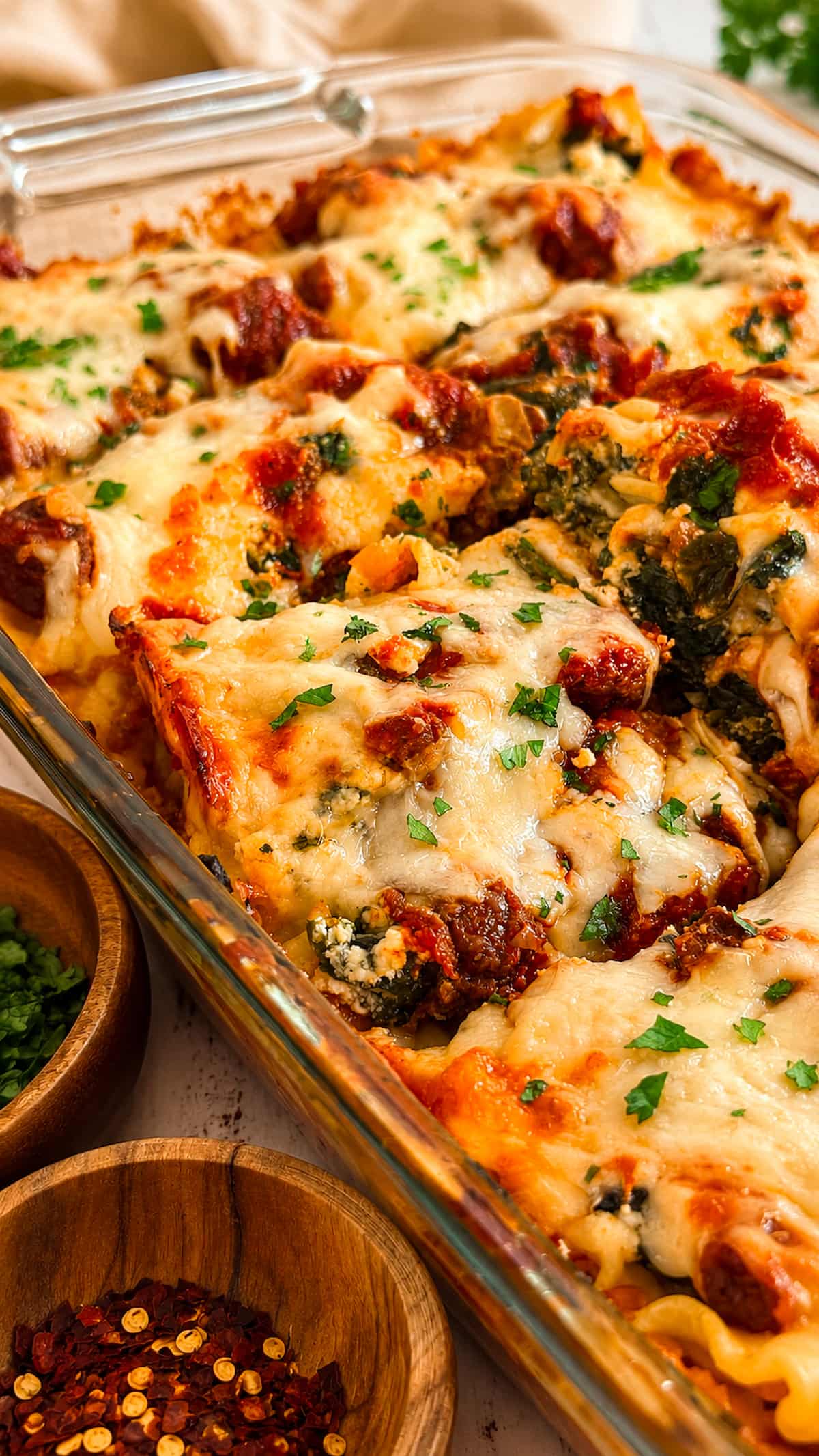 Close up of spinach and ground beef lasagna in a glass baking dish, topped with melted cheese, savoury meat sauce, and fresh parsley, with bowls of herbs and red pepper flakes nearby.