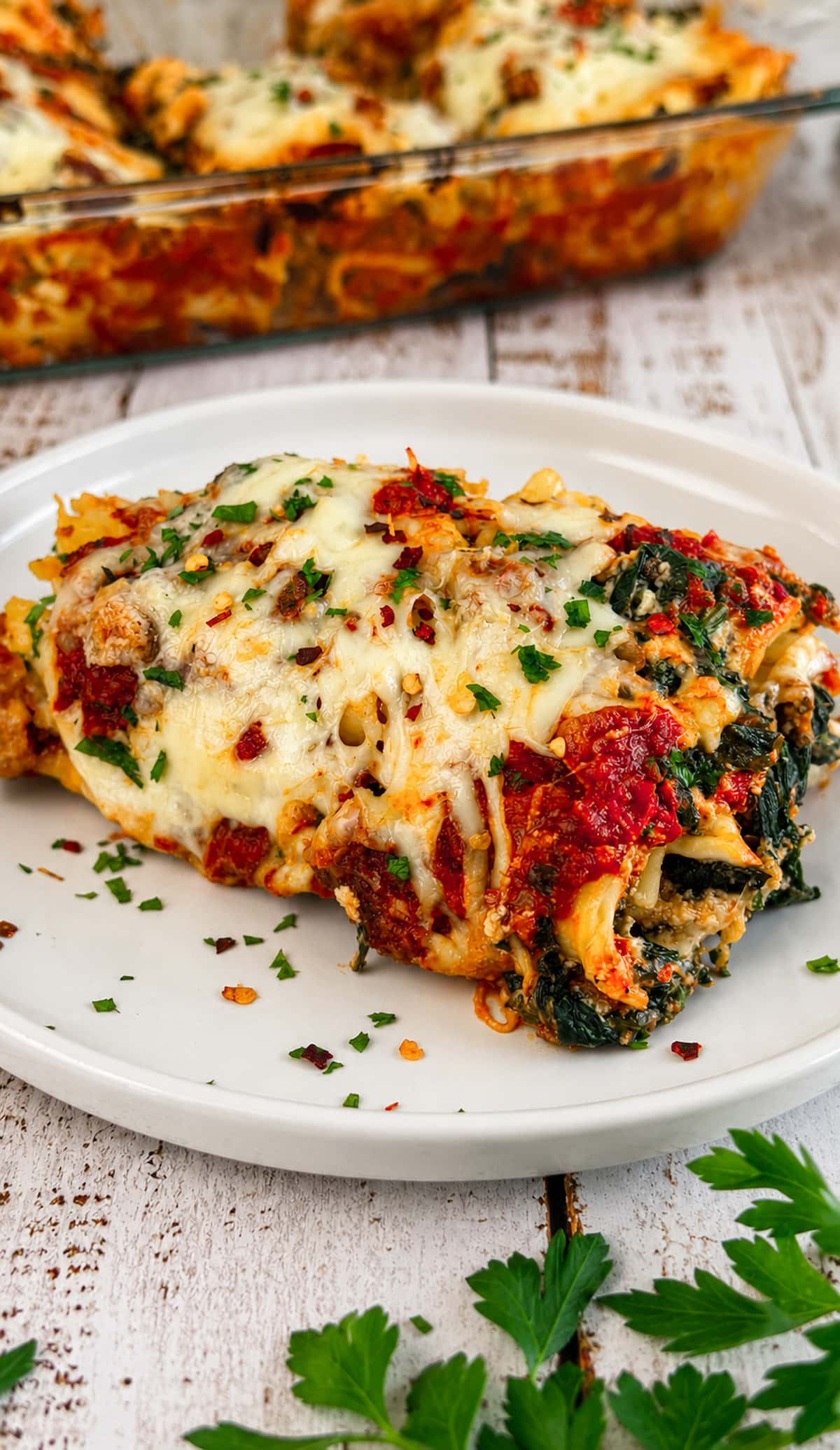 Slice of spinach and ground beef lasagna topped with melted cheese, parsley, and red pepper flakes on a white plate, with the baking dish blurred in the background.