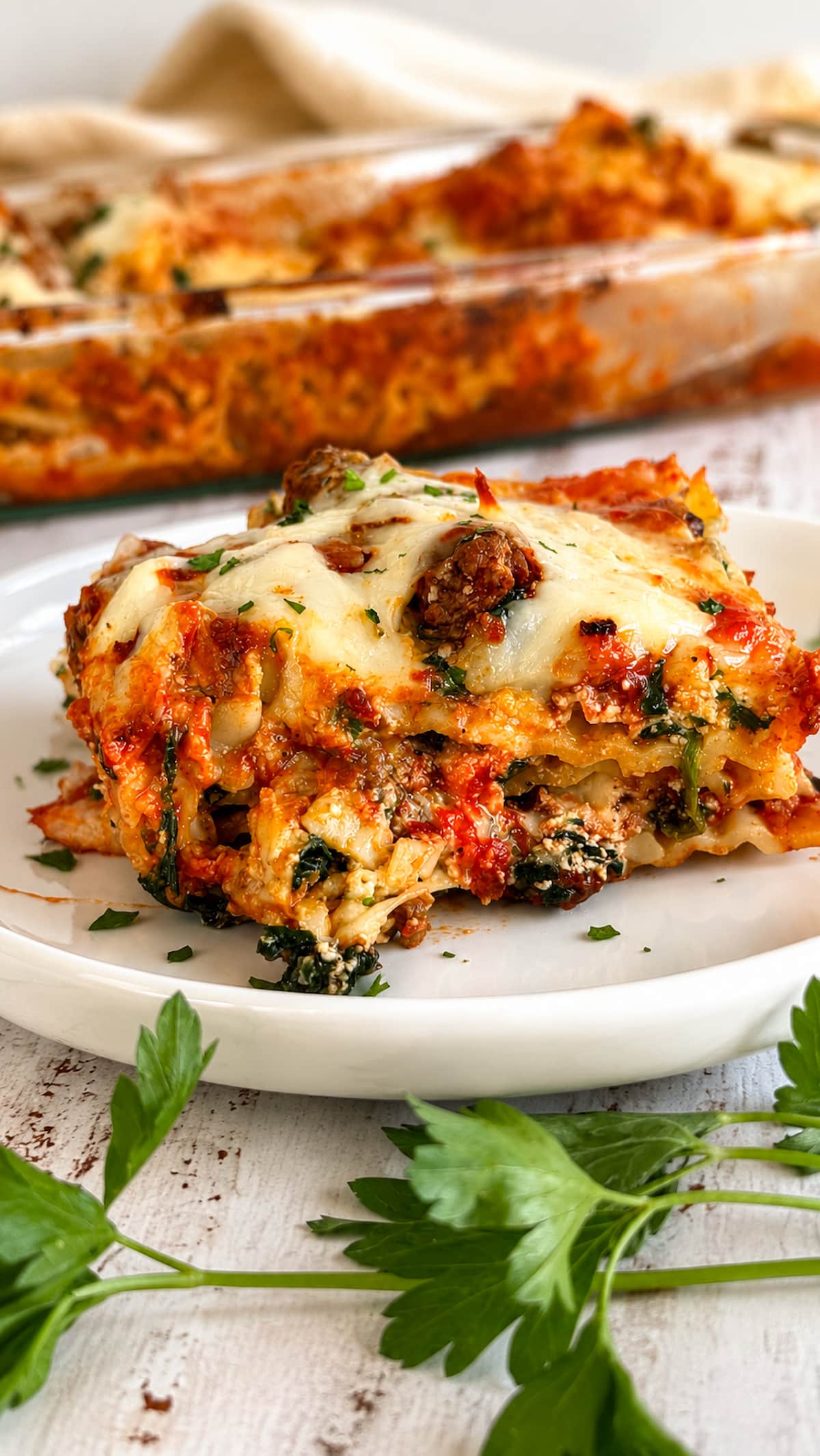Close up of a plated slice of spinach and ground beef lasagna with layers of pasta, rich meat sauce, spinach, and melted cheese, served with the baking dish blurred in the background.