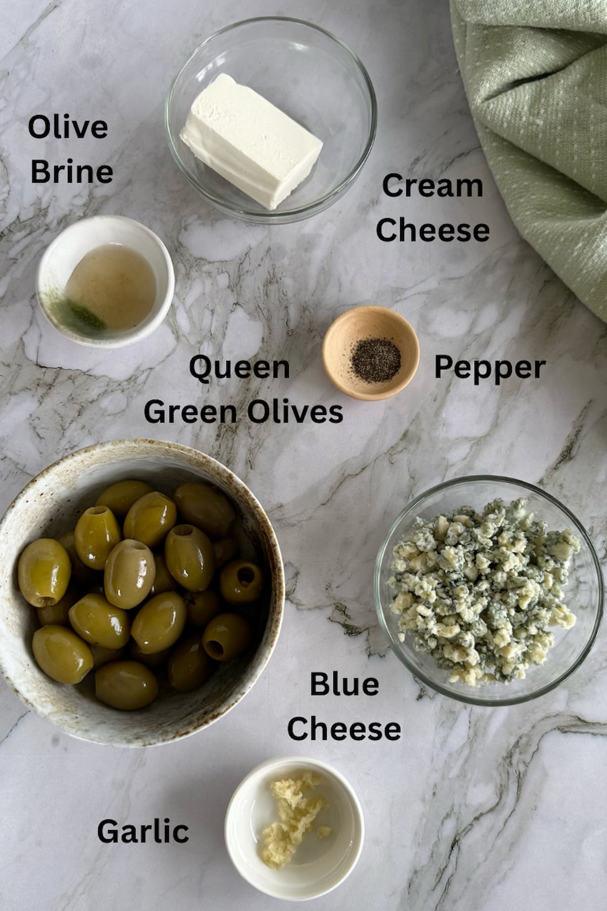 ingredients for blue cheese stuffed olives laid out in separate bowls - olive brine, cream cheese, queen green olives, pepper, blue cheese crumbles, and garlic.