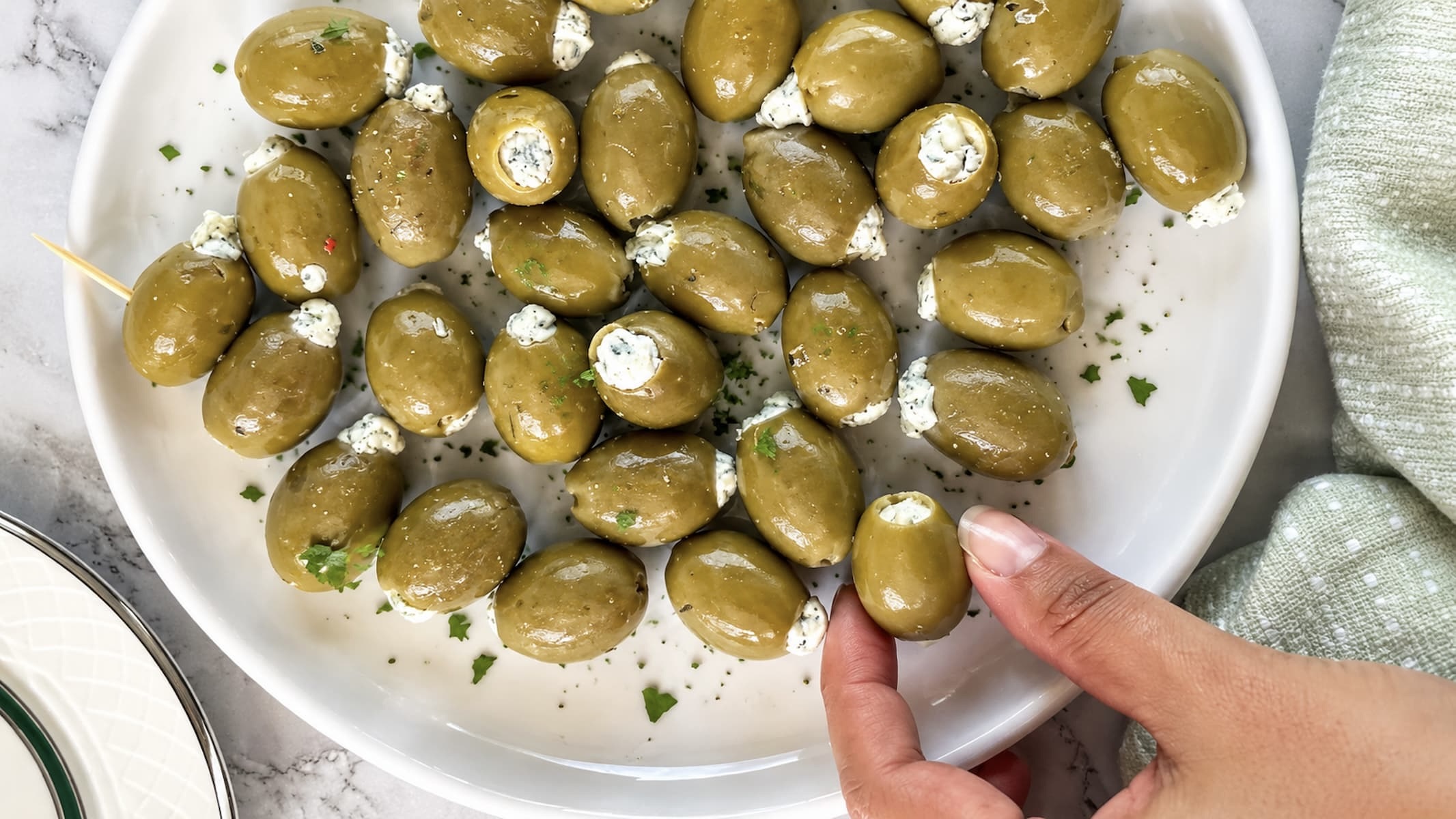 Overhead view of blue cheese stuffed green olives on a white plate, with a hand reaching for one olive and fresh parsley scattered on top.