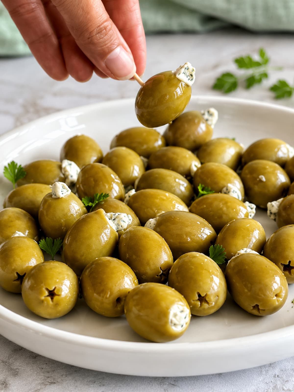 Hand picking up a blue cheese stuffed green olive with a toothpick above a plate with more garnished with fresh parsley.