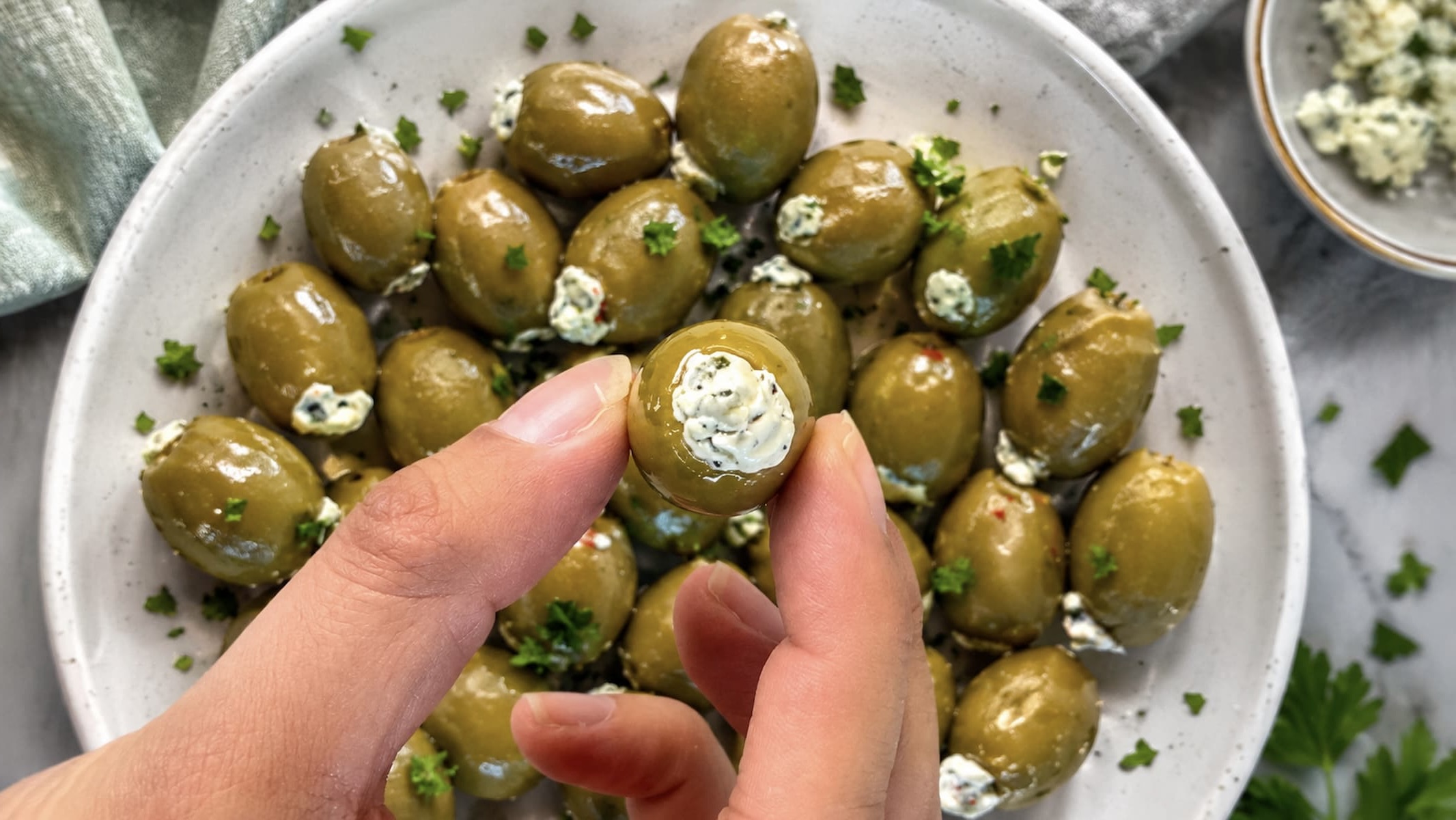 Hand holding a blue cheese stuffed green olive over a bowl filled with glossy green olives stuffed with creamy blue cheese, garnished with fresh parsley.
