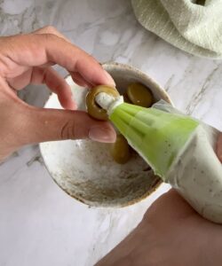 hand holding a queen green olive being stuffed with a creamy blue cheese mixture from a piping bag over a bowl with more olives.