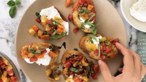 Hand grabbing burrata bruschetta made with creamy burrata cheese, colourful marinated cherry tomatoes, and fresh basil on toasted artisan bread.