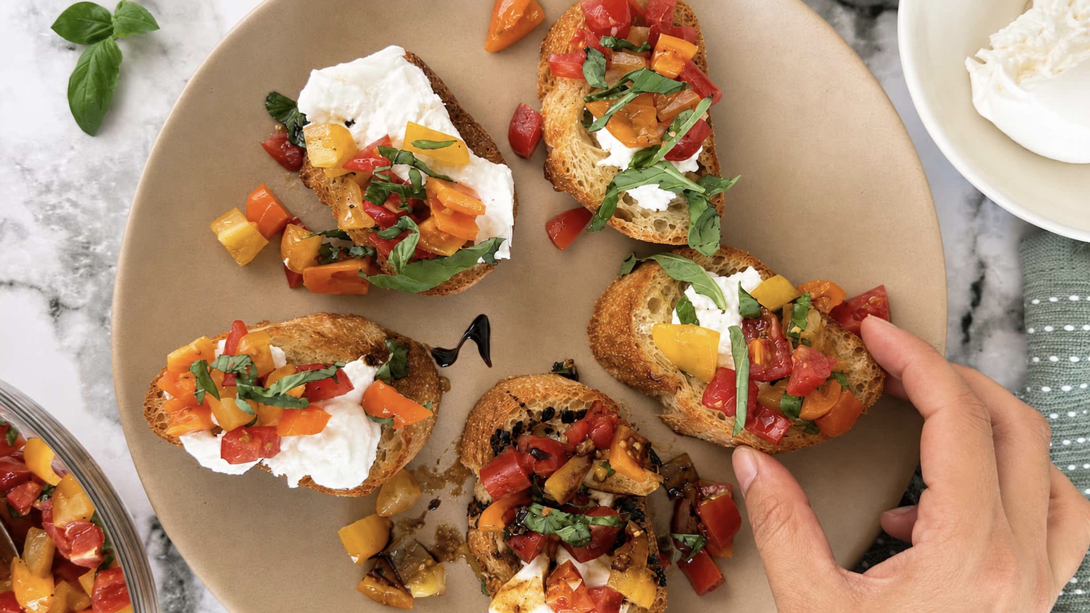 Hand grabbing burrata bruschetta made with creamy burrata cheese, colourful marinated cherry tomatoes, and fresh basil on toasted artisan bread.