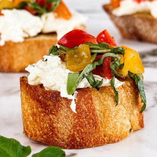 Burrata bruschetta made with creamy burrata cheese, marinated cherry tomatoes, and fresh basil on toasted artisan bread, photographed on a marble surface with a soft, blurred background.