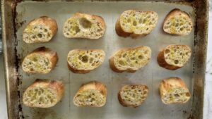 12 toasted pieces of artisan bread drizzled with olive oil laid out in a single layer on a baking sheet.
