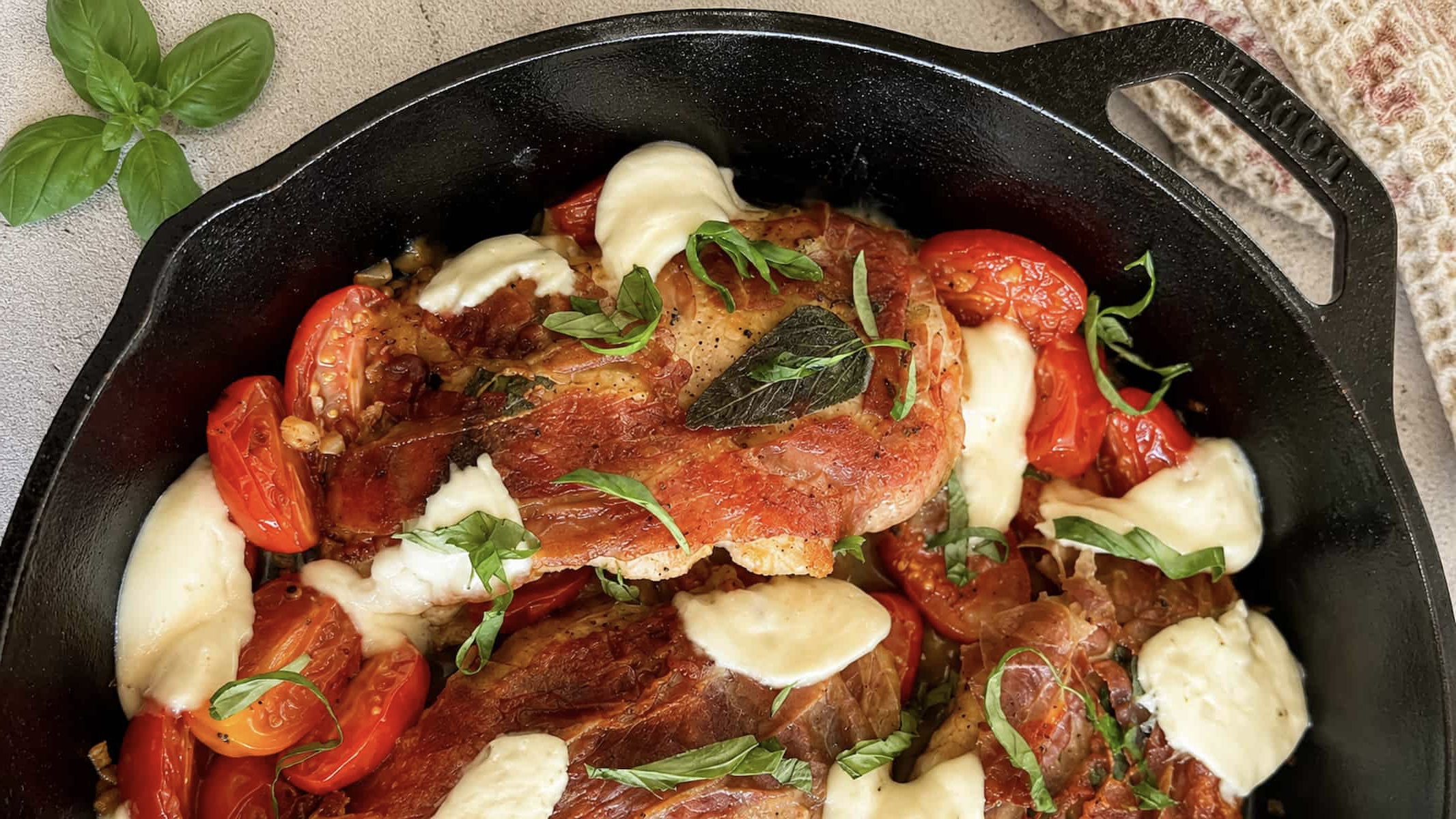 Close up of caprese chicken saltimbocca in a cast iron skillet with prosciutto wrapped chicken, melted mozzarella, roasted cherry tomatoes, and fresh basil.
