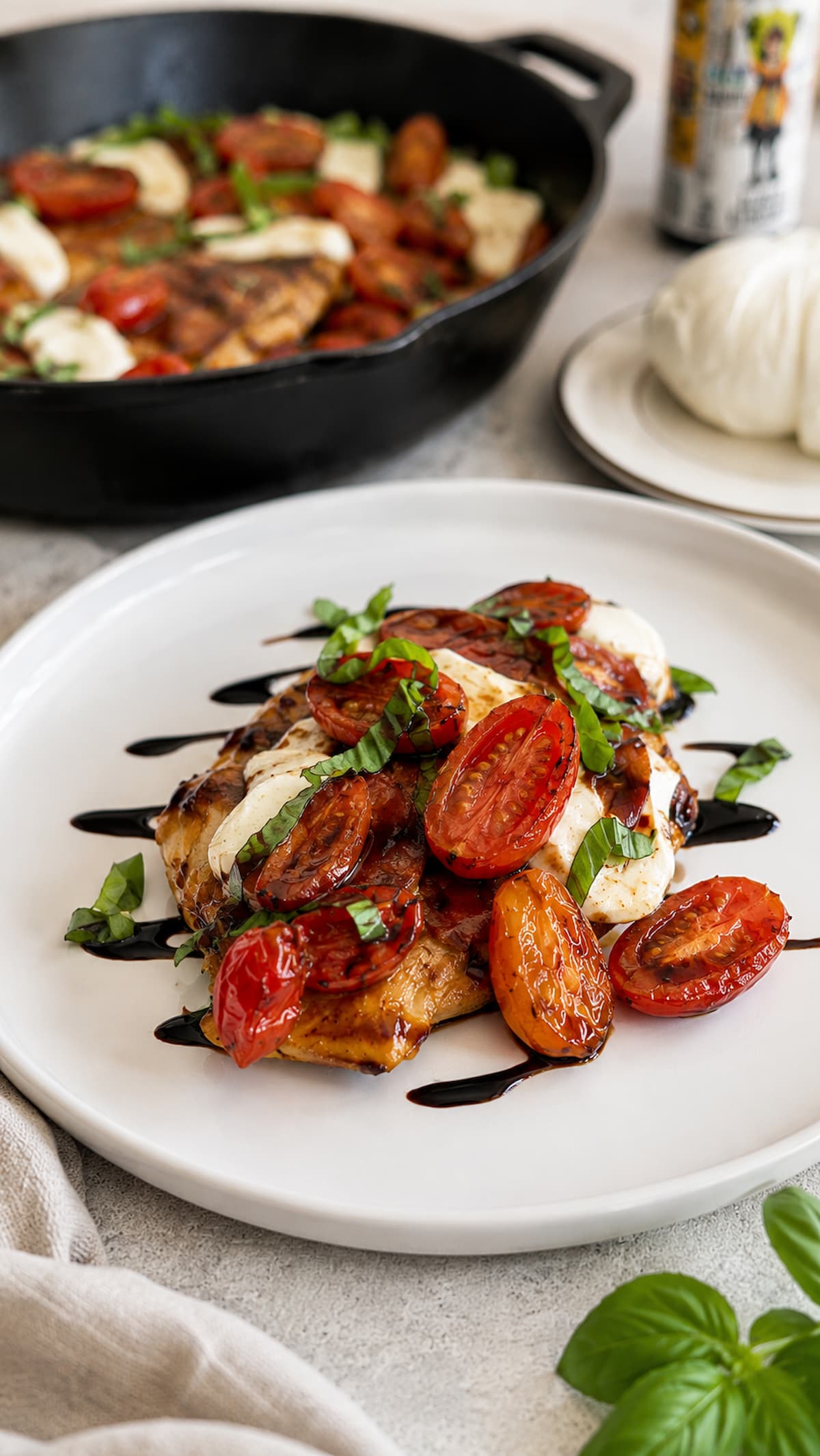 Plated caprese chicken saltimbocca topped with melted mozzarella, roasted cherry tomatoes, fresh basil, and balsamic glaze, with a cast iron skillet with more in the background.