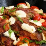 Close up of caprese chicken saltimbocca in a cast iron skillet with prosciutto wrapped chicken, melted mozzarella, roasted cherry tomatoes, and fresh basil.
