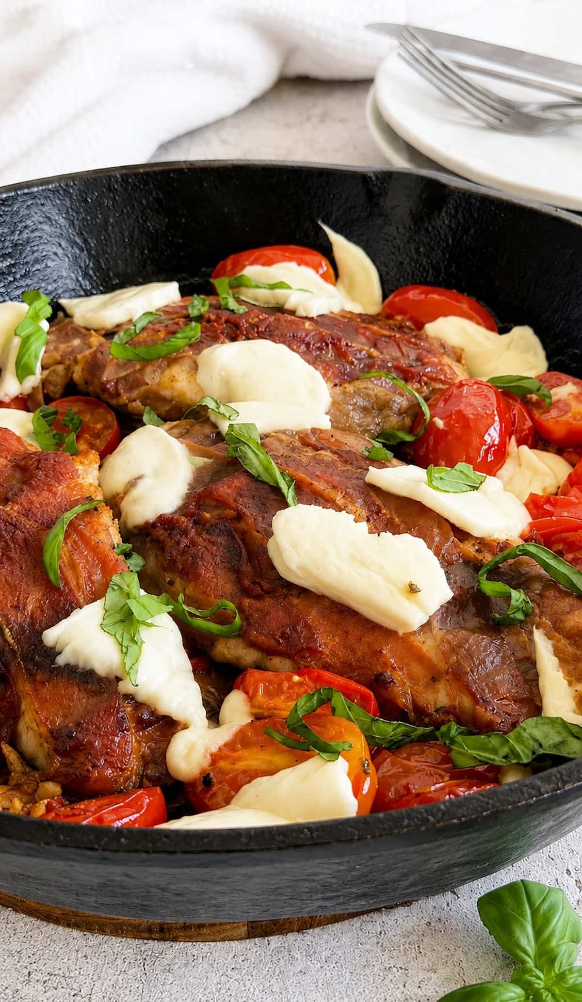 Close up of caprese chicken saltimbocca in a cast iron skillet with prosciutto wrapped chicken, melted mozzarella, roasted cherry tomatoes, and fresh basil.