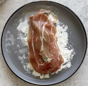 chicken breast topped with sage and prosciutto on a bed of flour on a grey plate.