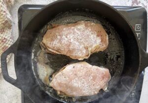 two uncooked floured and stuffed chicken breasts cooking in butter in a cast iron skillet on an induction cooktop.
