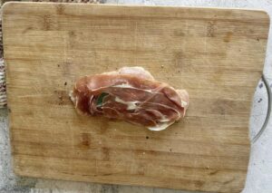 1 piece of raw chicken topped with prosciutto and sage on a cutting board.