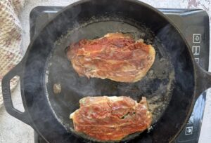 two crispy prosciutto topped chicken breasts cooking in a cast iron skillet on an induction burner.