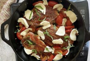 caprese chicken saltimbocca in a skillet with prosciutto wrapped chicken breasts, torn fresh mozzarella pieces, cherry tomatoes, and fresh basil.