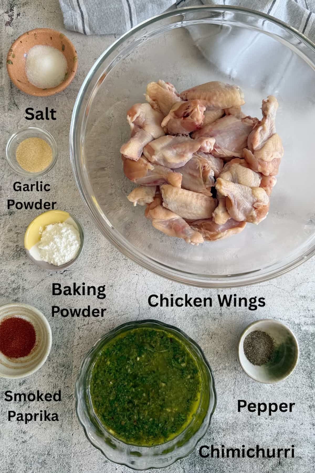 ingredients for chimichurri wings laid out in separate bowls - chicken wings, chimichurri, baking powder, salt, pepper, garlic powder, and smoked paprika.