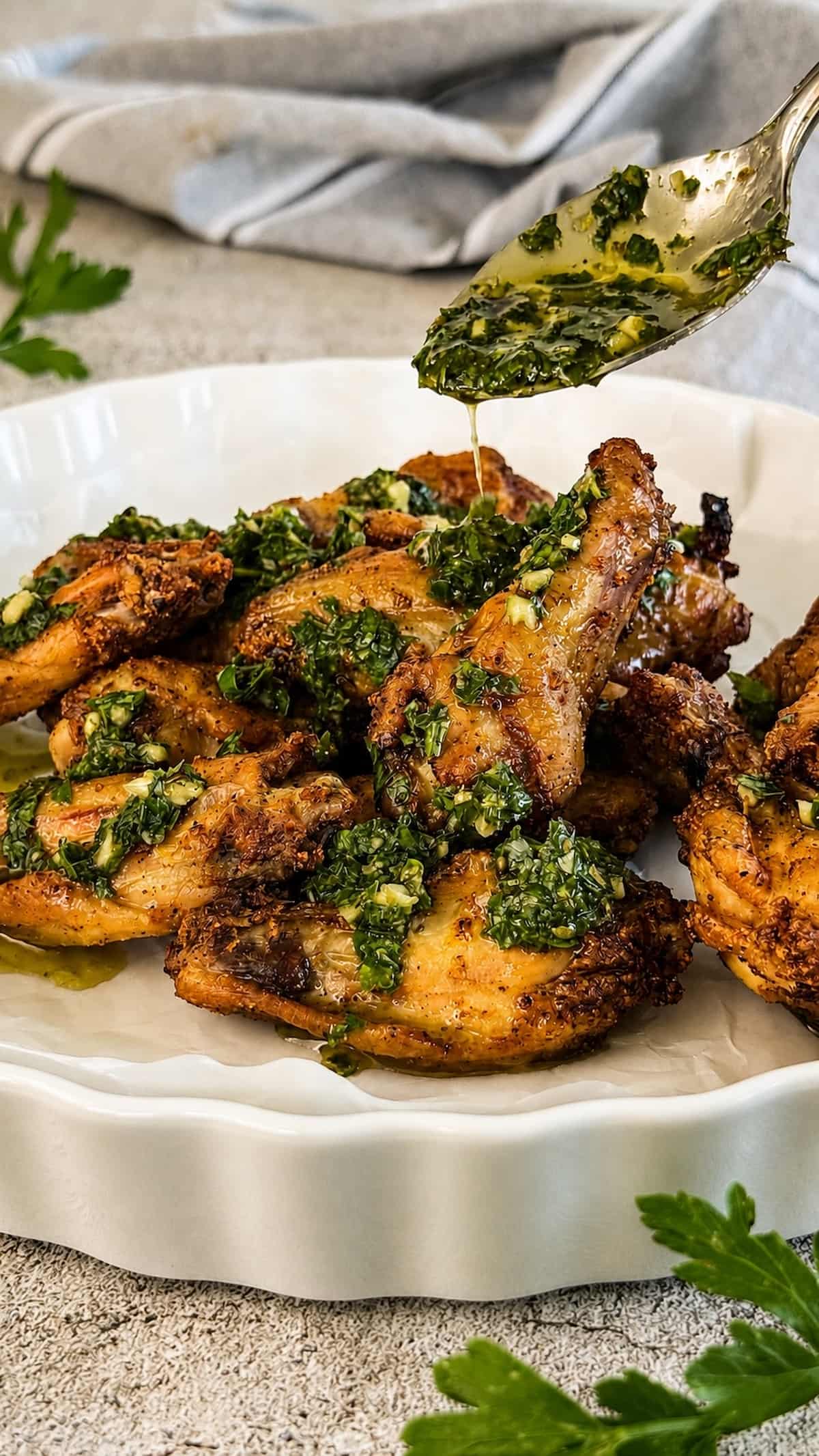 Close up of crispy chimichurri chicken wings in a white serving dish as fresh chimichurri sauce is spooned over the top.