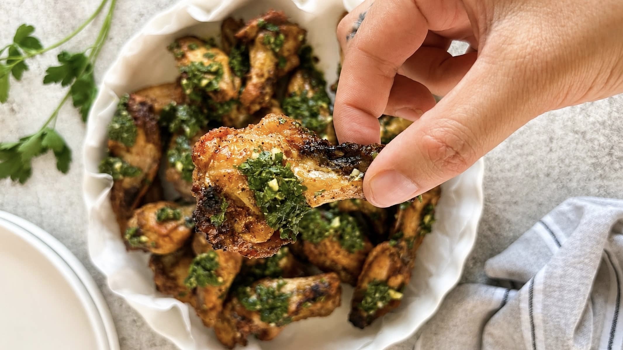 Hand holding a crispy chimichurri chicken wing in front of a bowl of golden brown wings topped with fresh herb sauce.
