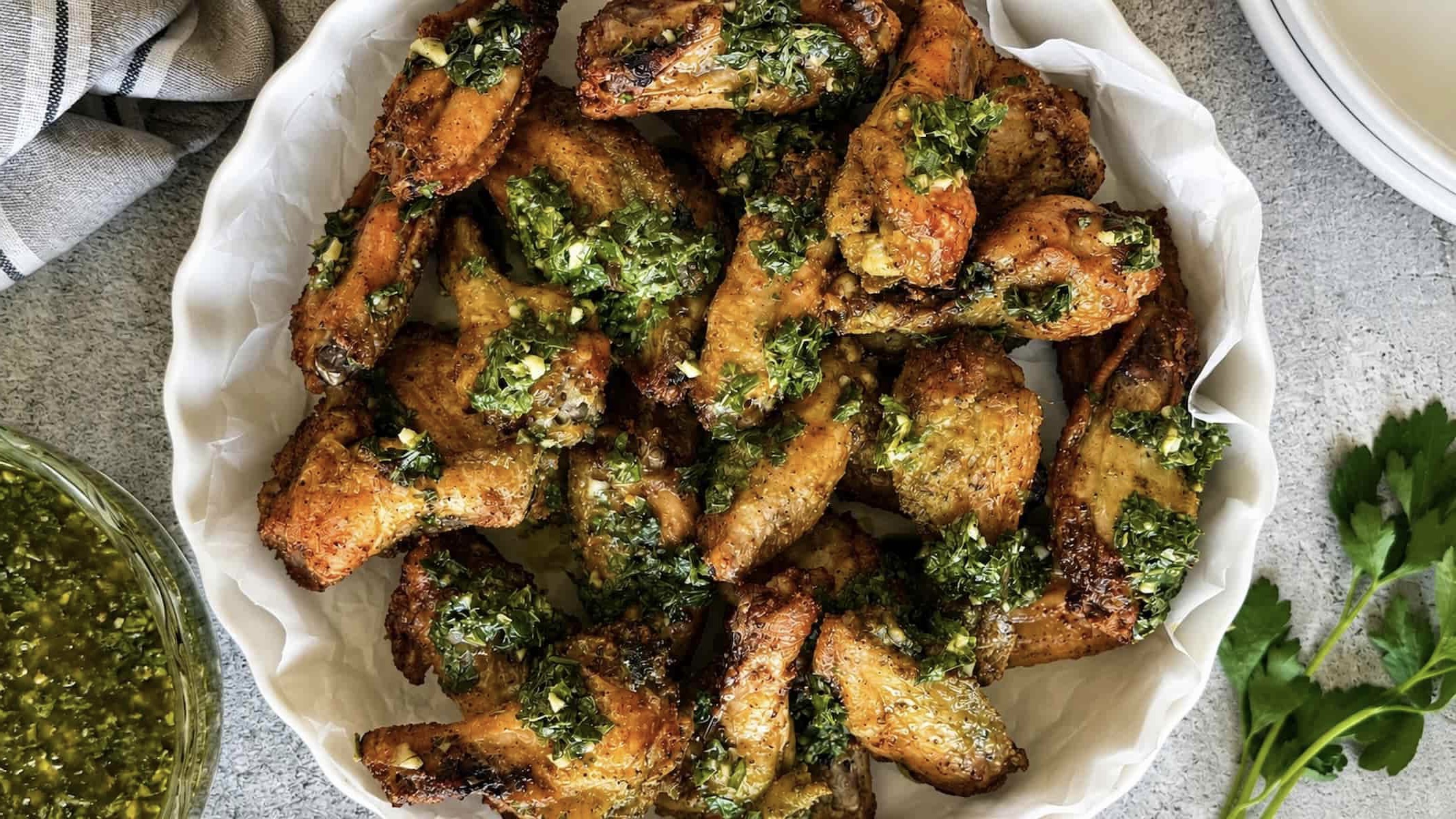 Overhead view of golden brown chimichurri chicken wings piled in a white serving dish with fresh parsley and chimichurri sauce nearby.