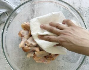 hand with a paper towel patting raw chicken wings down in a large glass bowl.