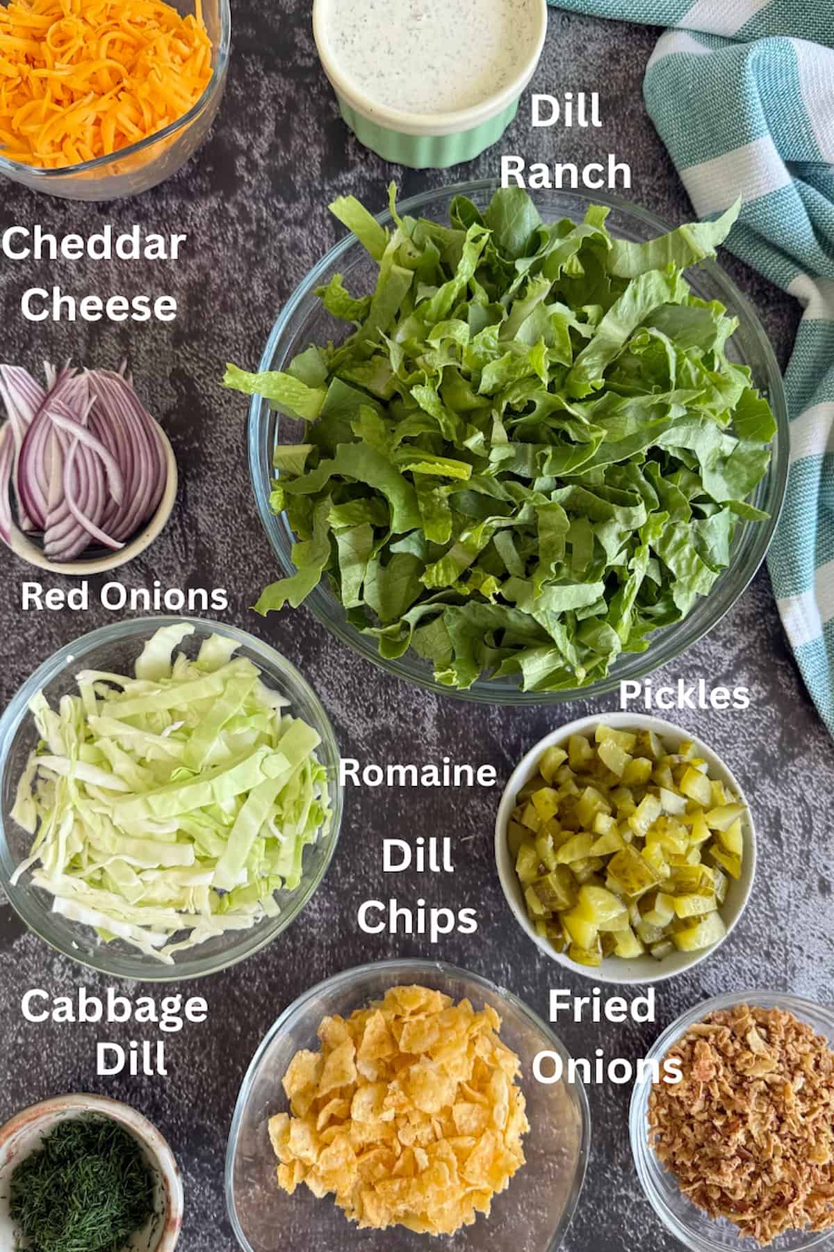 ingredients to make dill pickle chopped salad laid out in separate bowls - romaine, cabbage, red onions, fried onions, cheddar cheese, dill ranch, fresh dill, and dill pickle chips.