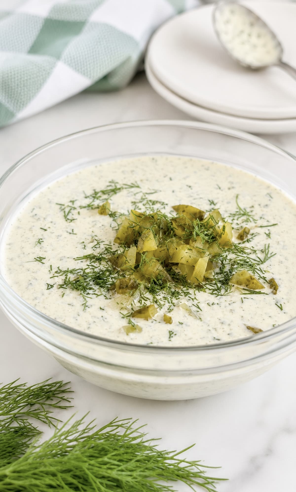 Creamy homemade dill pickle ranch dressing garnished with chopped pickles and fresh dill in a glass bowl, fresh dill in the front, two plates with a spoon with dressing on it in the back.
