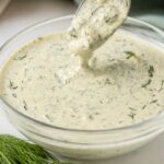 Creamy homemade dill pickle ranch dressing in a glass bowl with a spoonful lifted above, garnished with fresh dill on a marble countertop.