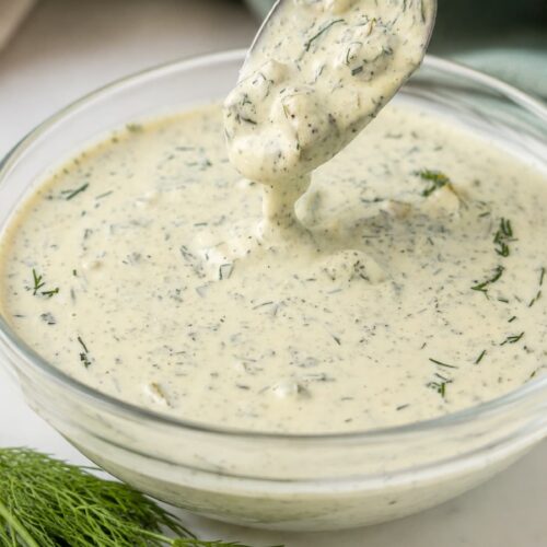 Creamy homemade dill pickle ranch dressing in a glass bowl with a spoonful lifted above, garnished with fresh dill on a marble countertop.
