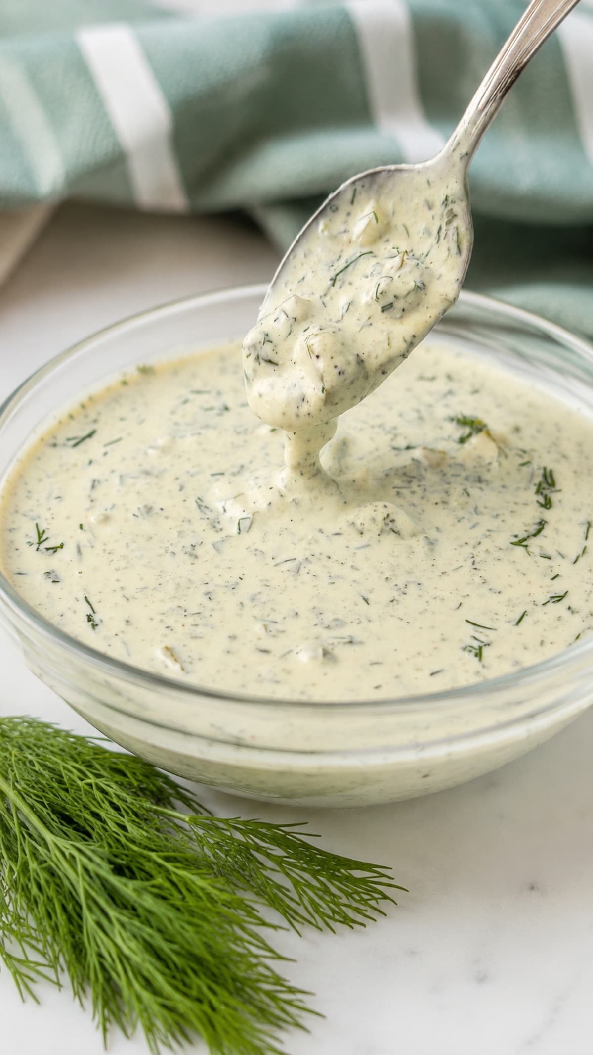 Creamy homemade dill pickle ranch dressing in a glass bowl with a spoonful lifted above, garnished with fresh dill on a marble countertop.