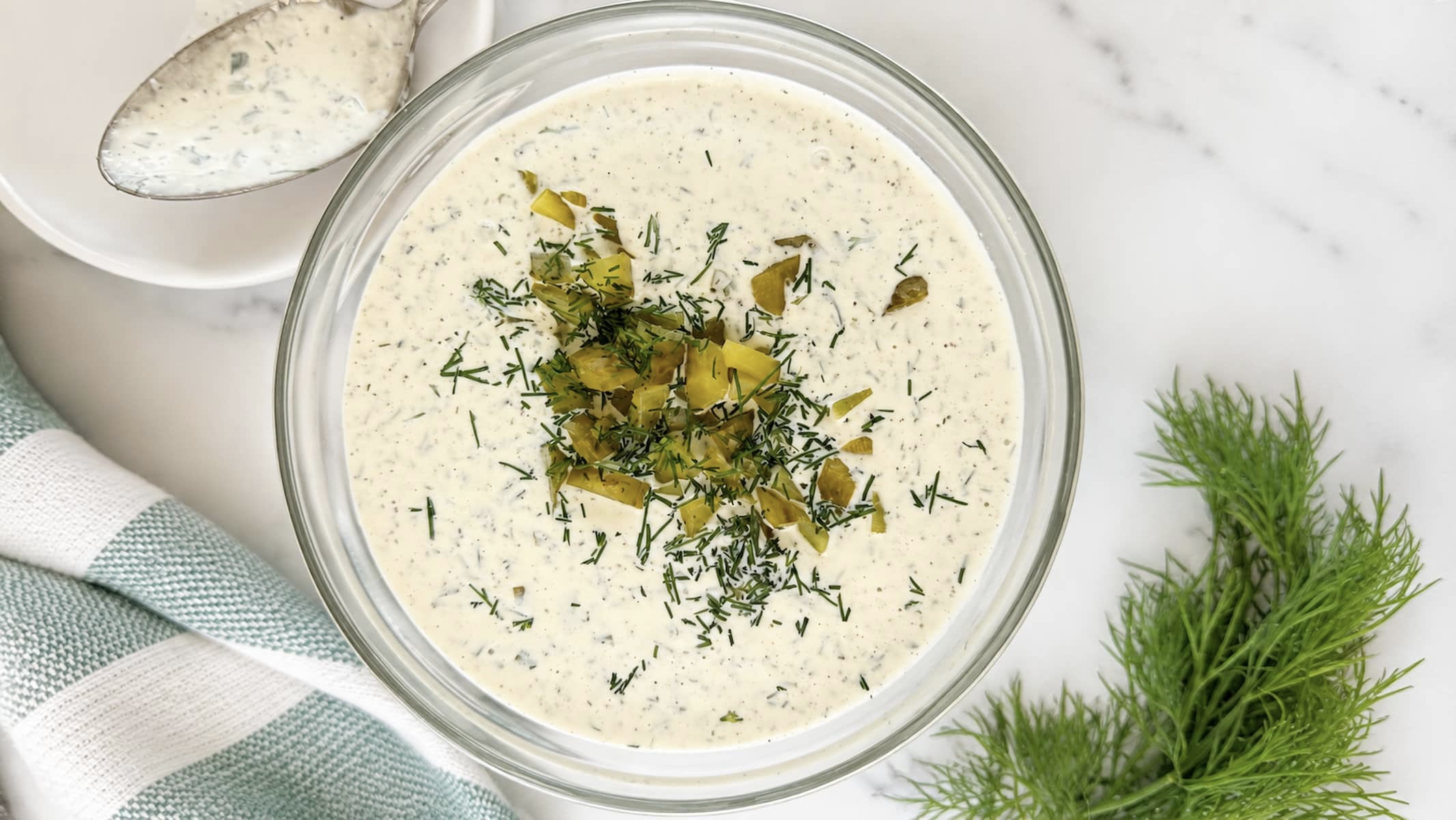 Top-down view of homemade dill pickle ranch dressing garnished with chopped pickles and fresh dill in a glass bowl. A spoon rests on a white plate beside the bowl, with fresh dill sprigs.