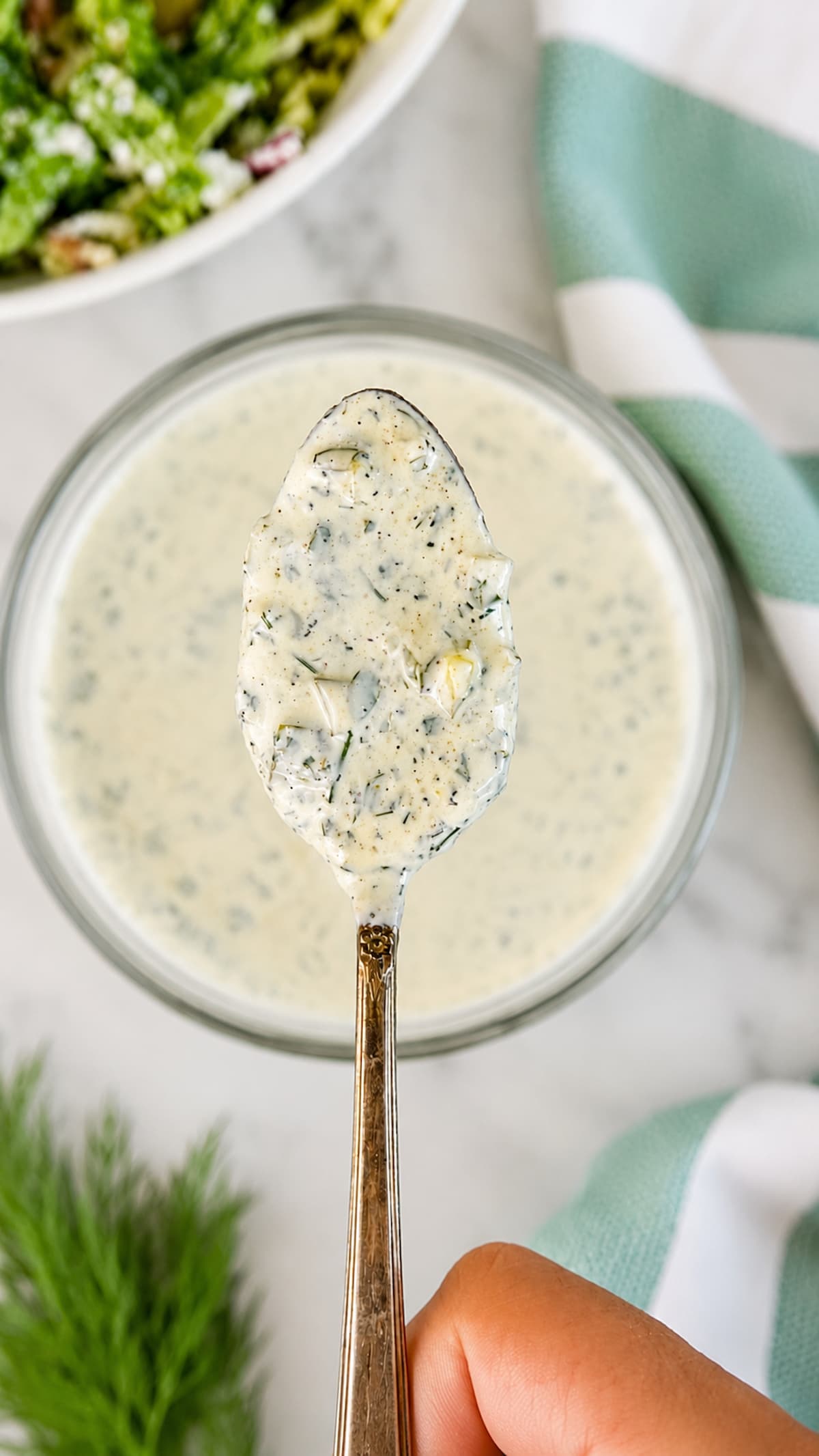 Hand holding a spoonful of homemade dill pickle ranch dressing above a bowl of creamy dill pickle ranch.