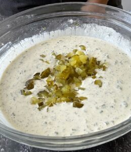 glass bowl of creamy ranch dressing with chopped pickles on top.