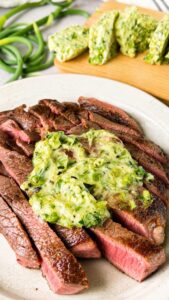 Garlic scape butter melting over sliced medium rare steak with extra garlic scape butter on a wooden board in the background.