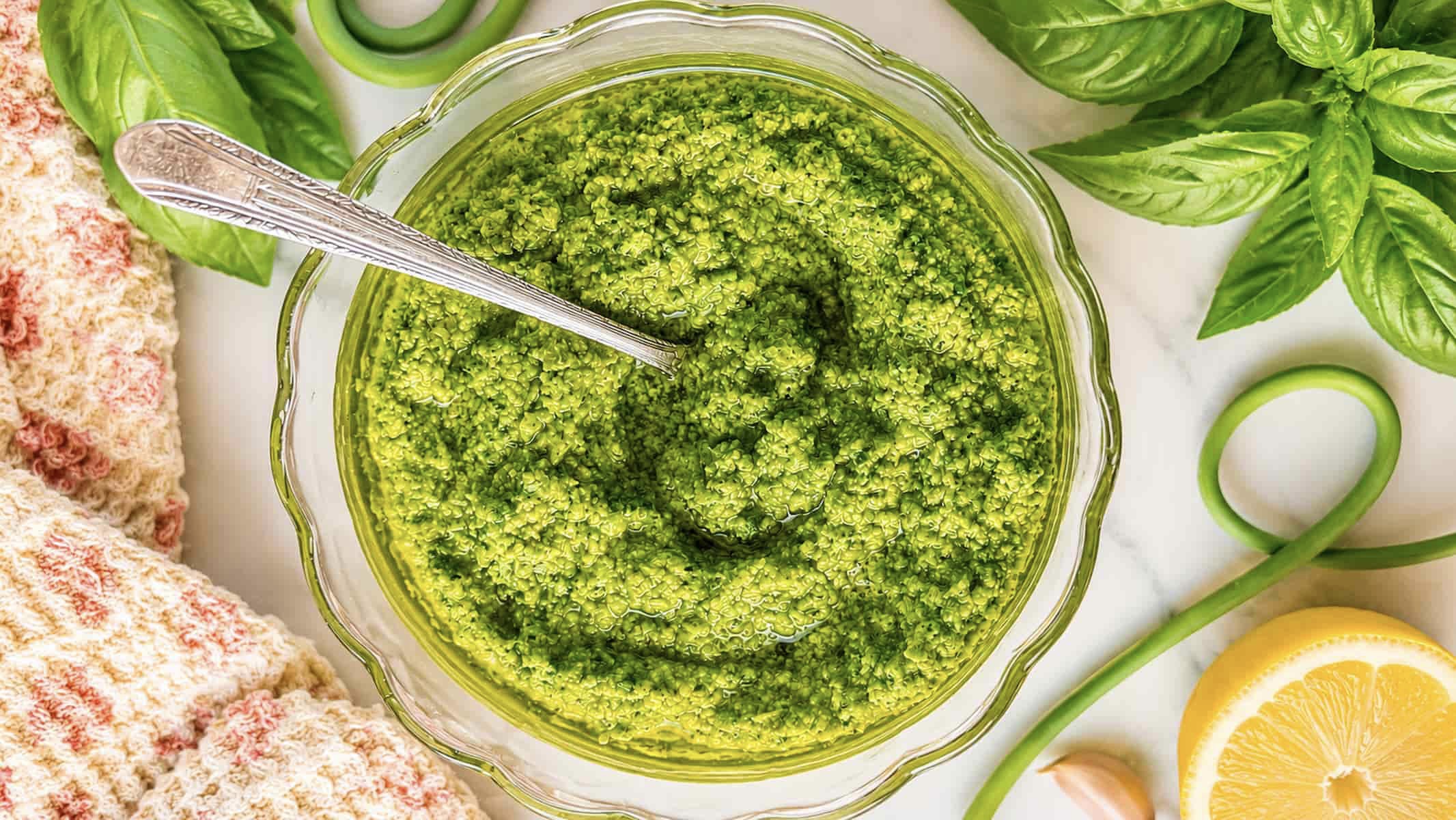 Homemade garlic scape pesto in a glass bowl garnished with olive oil, fresh basil, lemon, and garlic scapes.