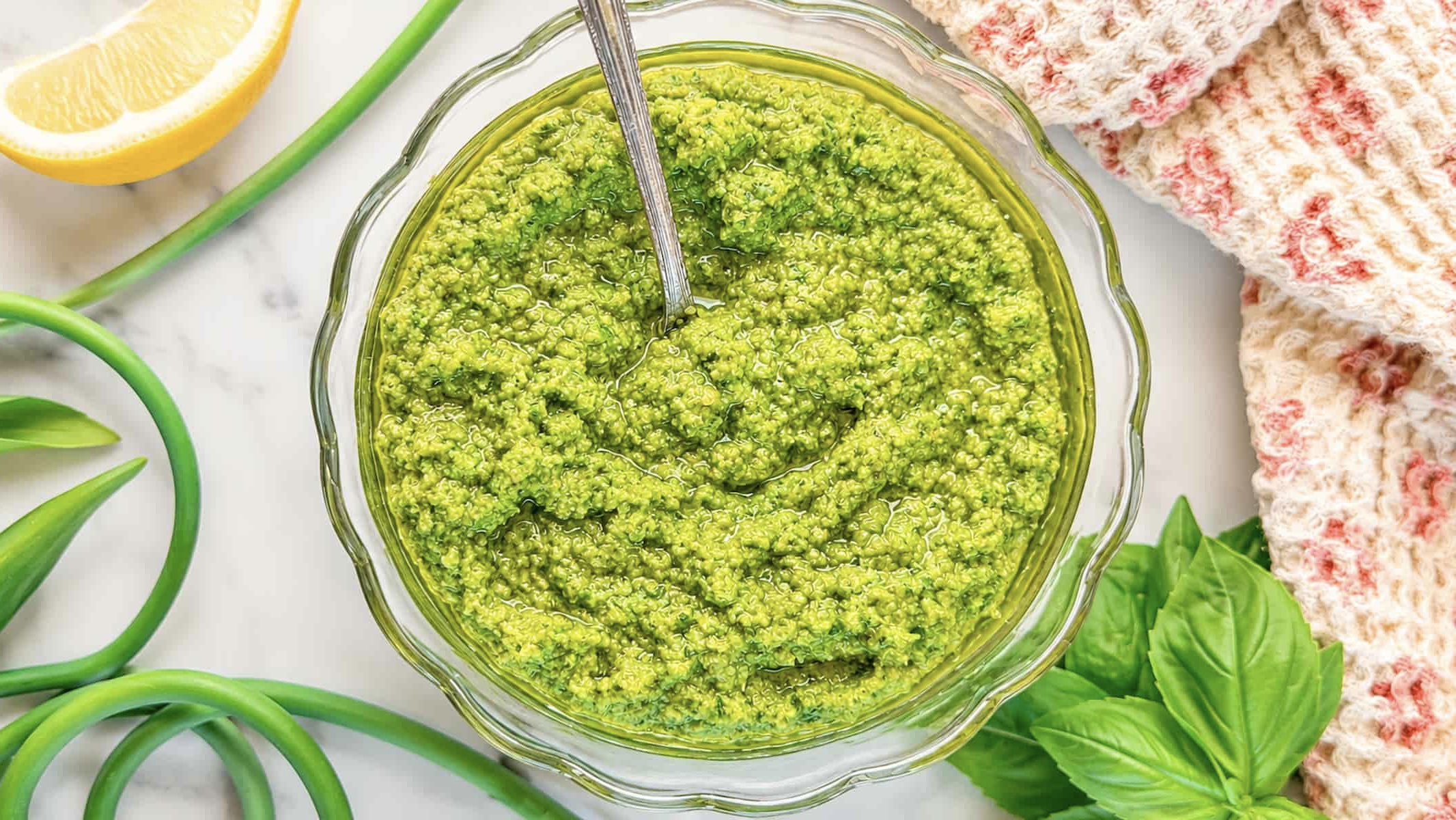 Garlic scape pesto in a glass bowl with basil leaves, lemon, and fresh garlic scapes.