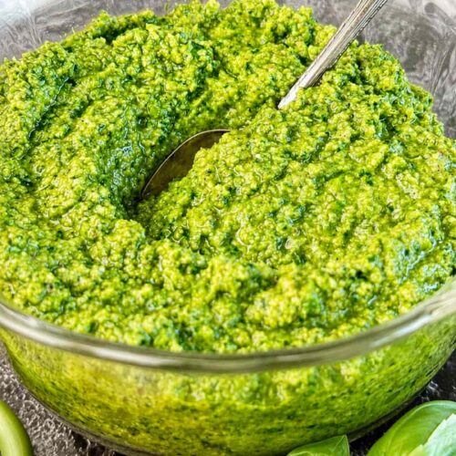 Close up of homemade garlic scape pesto in a glass bowl with fresh basil leaves and a lemon wedge.
