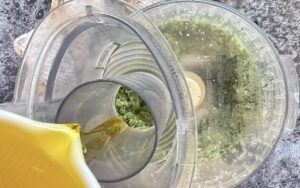 olive oil being drizzled into the chute of a food processor making garlic scape pesto.