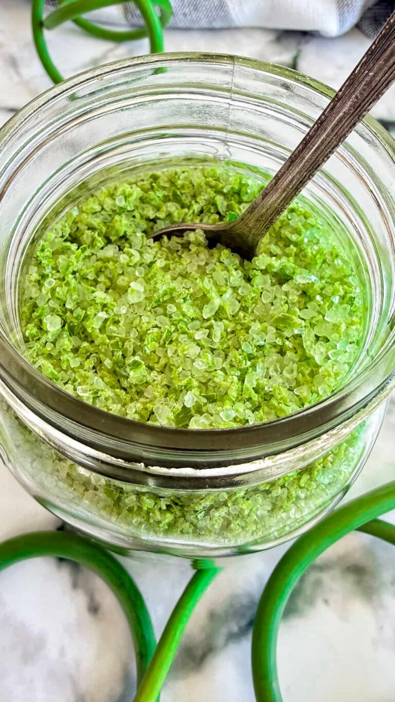 Garlic scape salt in a glass jar with coarse sea salt crystals and a small wooden spoon.