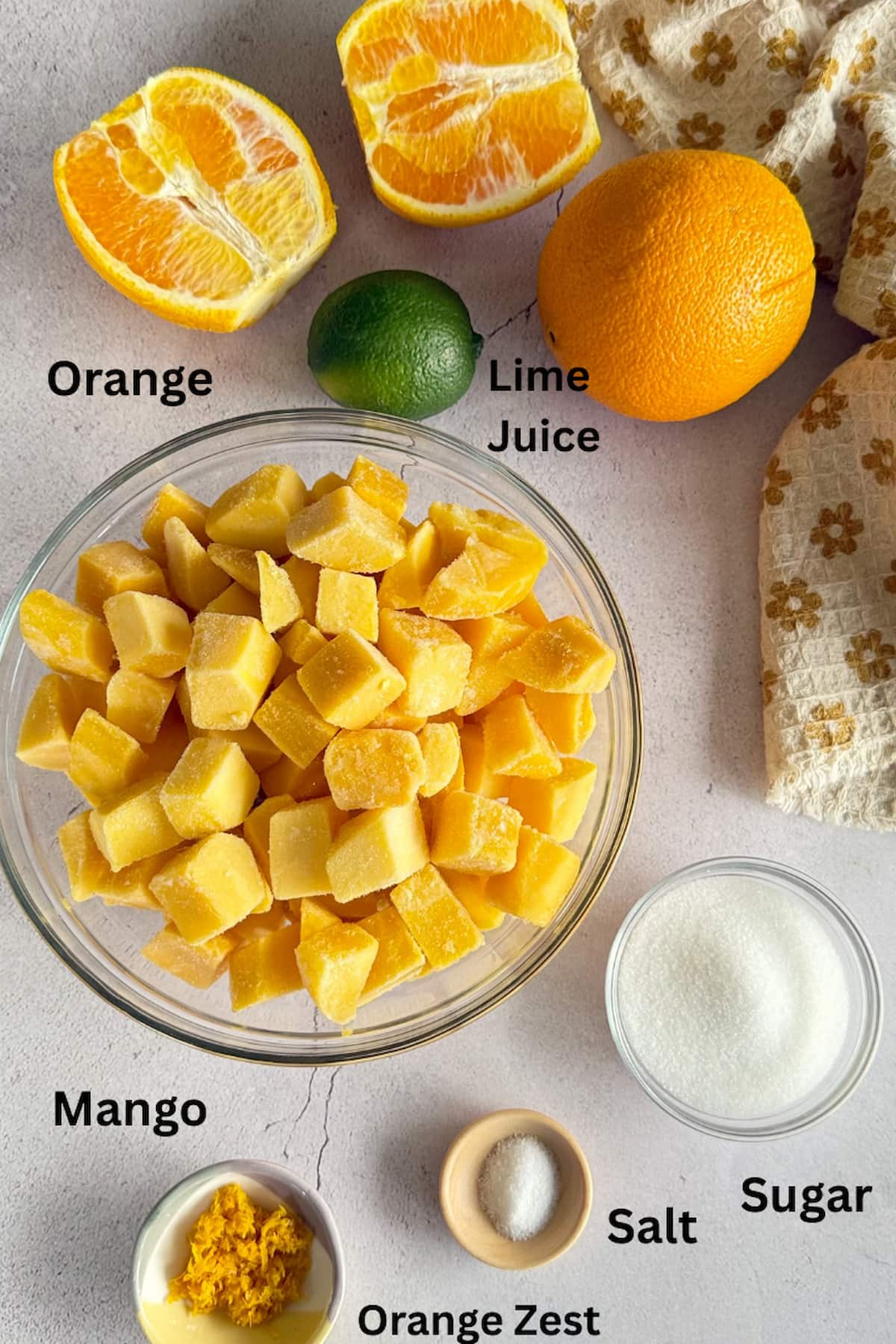 ingredients for mango orange sorbet laid out in bowls - frozen mango chunks, orange zest, salt, and sugar, next to a whole lime, whole orange, and halved orange.