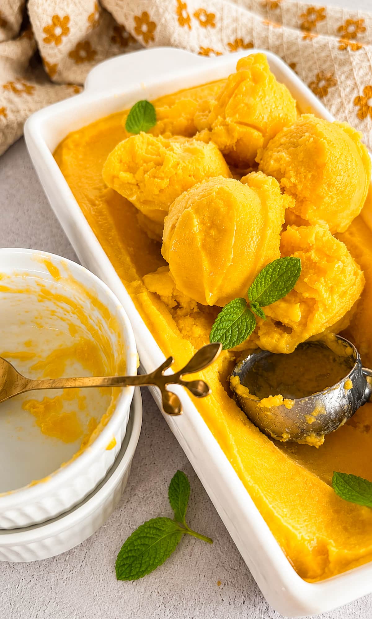 Mango orange sorbet scoops in a white baking dish with fresh mint, a metal ice cream scoop, and a serving bowl on a light stone surface.