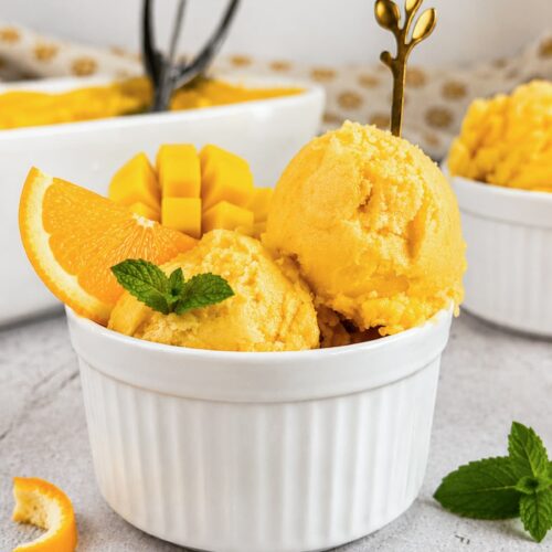 Mango orange sorbet scoops in a white ramekin with a fresh orange slice, diced mango, and mint garnish.