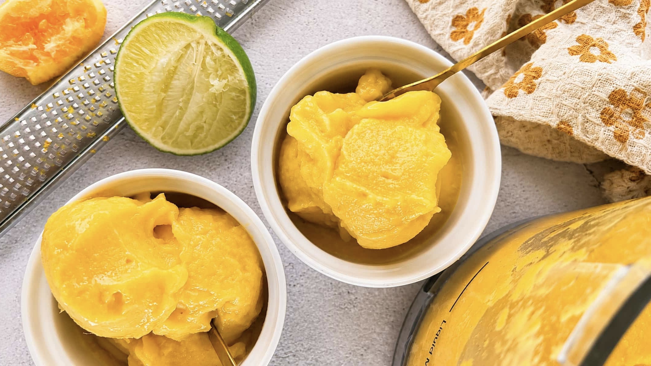 Mango orange sorbet scoops in white ramekins with fresh lime, orange, and a citrus zester on a light stone surface.