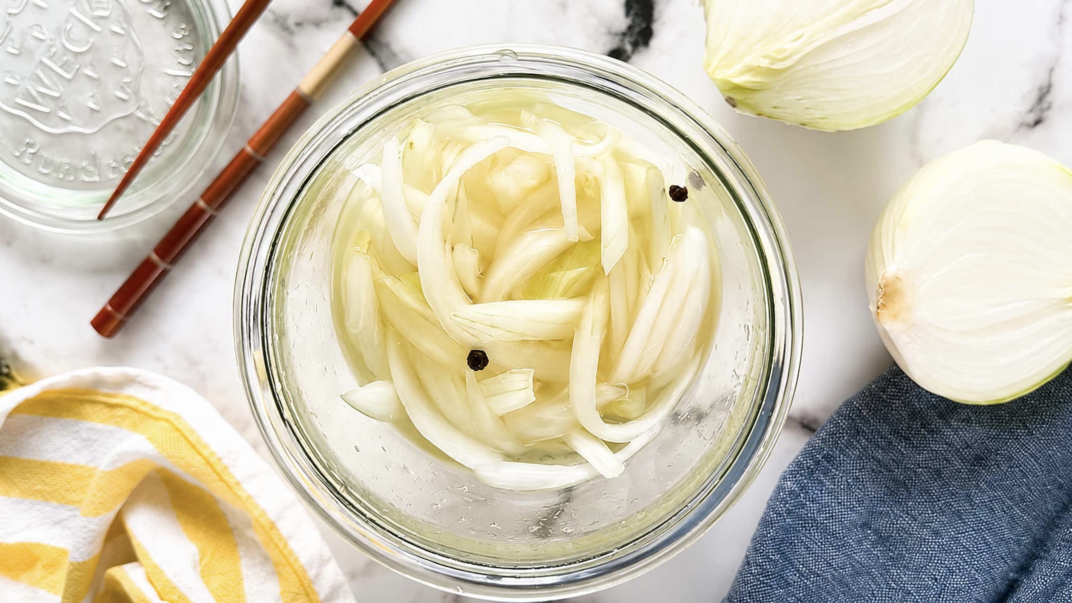 Pickled white onions in a glass jar with peppercorn brine, surrounded by fresh onion halves, chopsticks, and a striped kitchen towel.