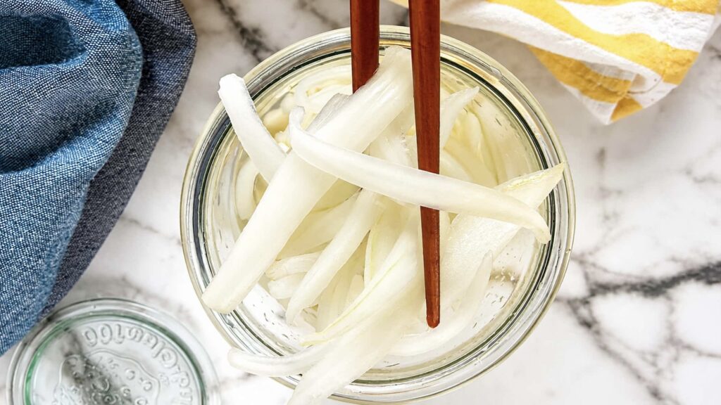Pickled white onions in a glass jar with onion slices lifted by chopsticks over a light brine.