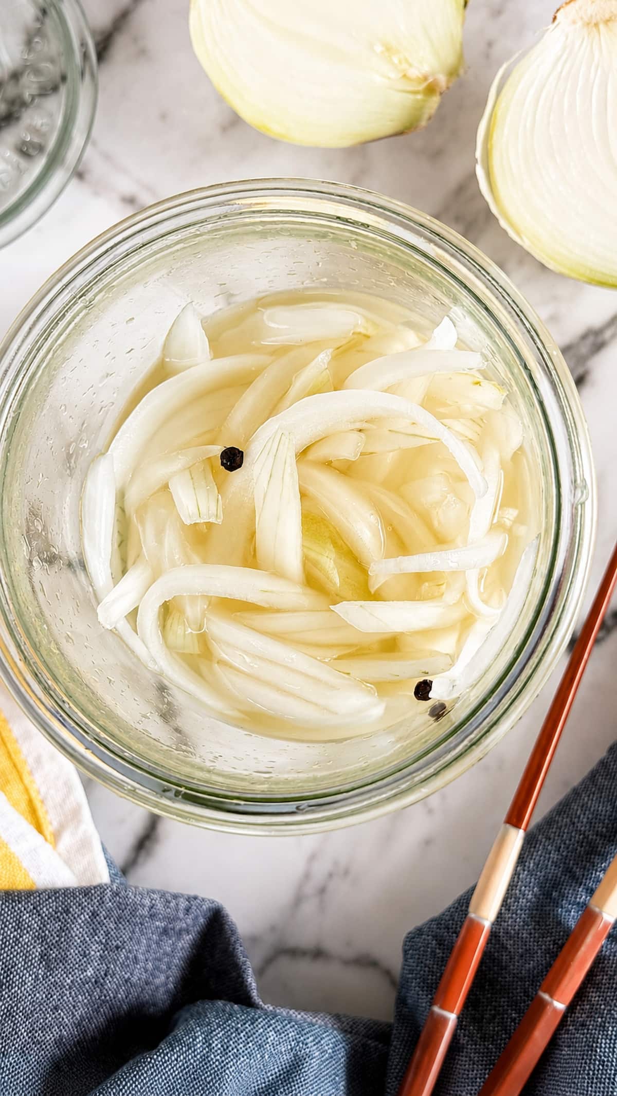 Pickled white onions in a glass jar with peppercorn brine, surrounded by fresh onion halves and chopsticks.